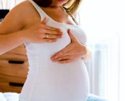 A woman in a white top holding her stomach as she lives to the journey of parenthood.